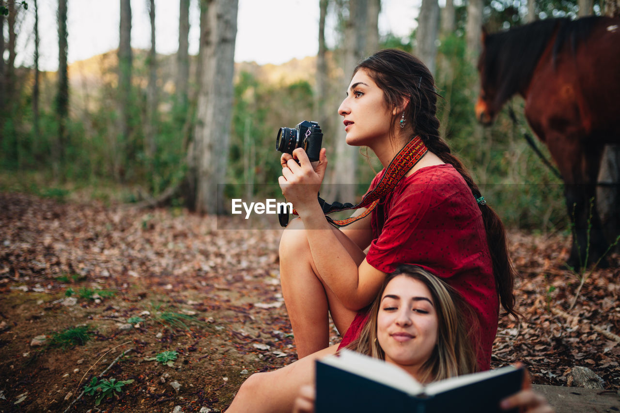 Woman reading book while friend photographing in forest