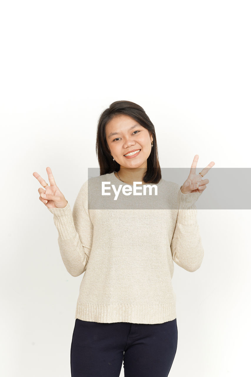 finger, smiling, one person, happiness, portrait, women, studio shot, gesturing, hand, adult, emotion, looking at camera, arm, white background, cheerful, indoors, front view, clothing, standing, cut out, female, sleeve, positive emotion, three quarter length, young adult, casual clothing, hand sign, communication, thumbs up, smile, hand raised, teeth, outerwear, joy, person, relaxation, copy space, showing, waist up, laughing, excitement, limb, t-shirt, hairstyle, enjoyment, lifestyles, sign language
