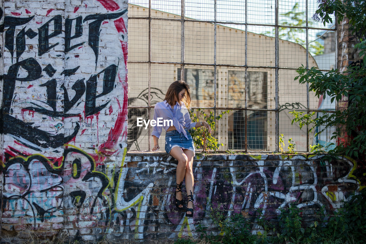 Young woman sitting on window sill amidst graffiti wall