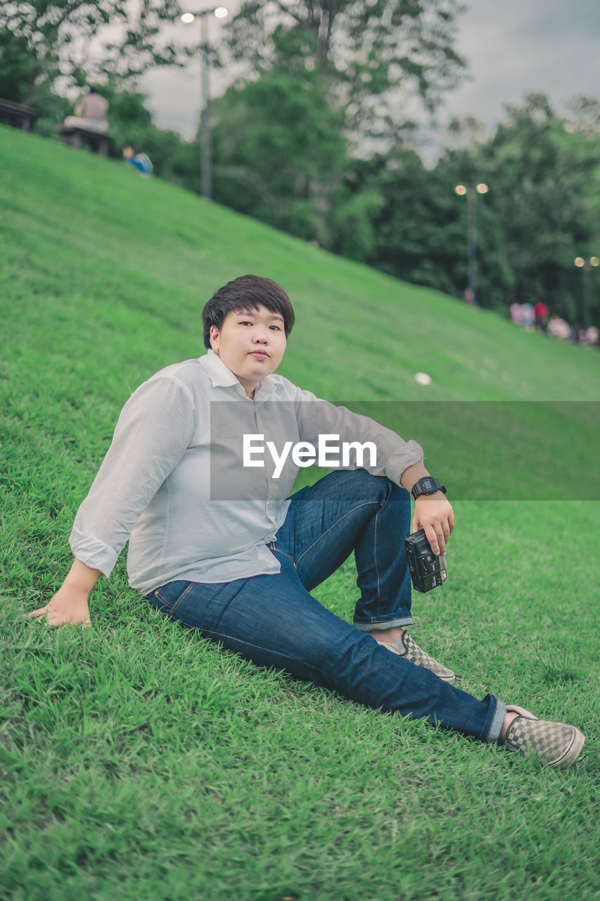 Portrait of young woman with camera sitting on grassy land in park