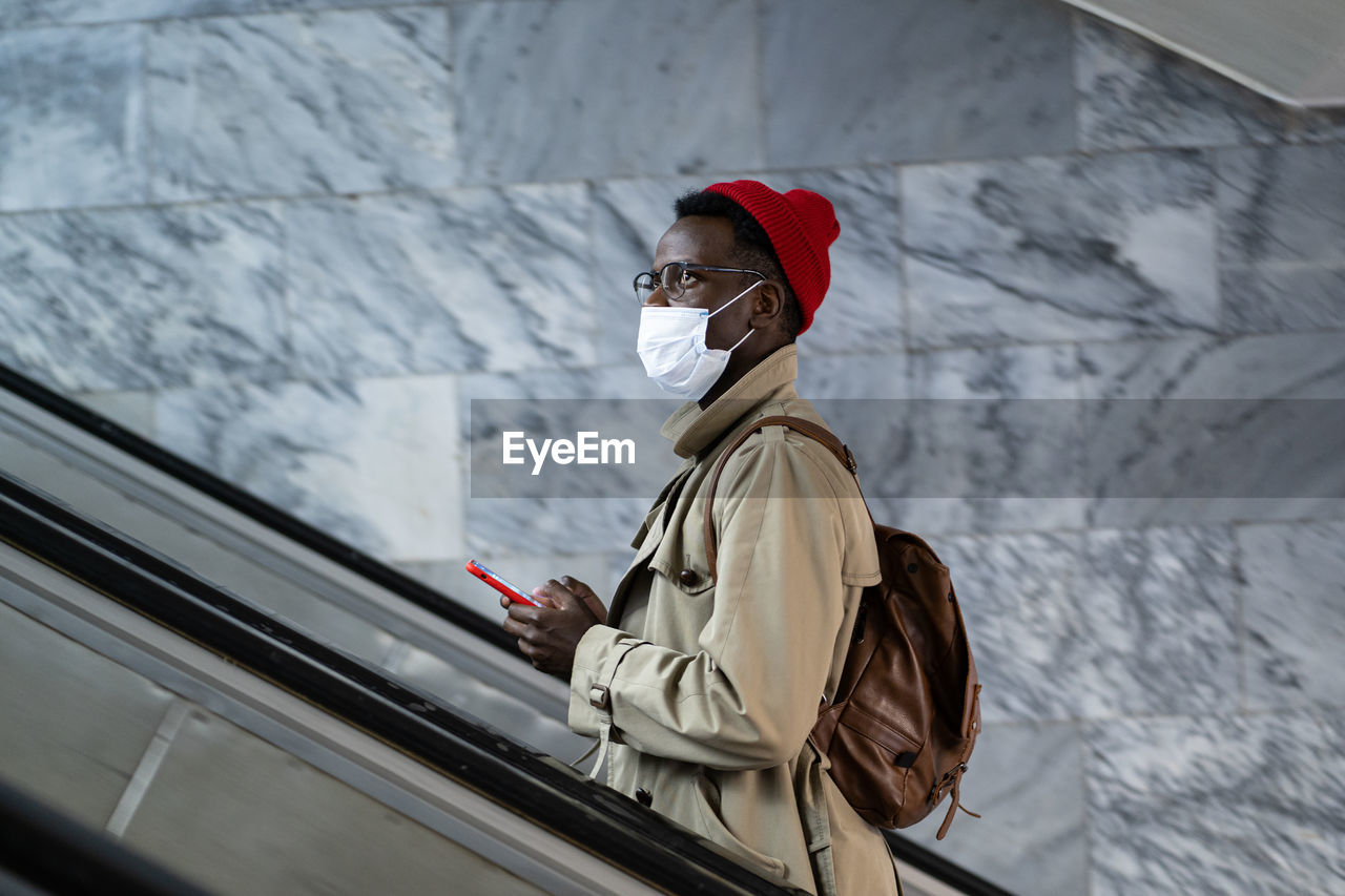 Pensive african american millennial man with smartphone on escalator subway wearing protective mask