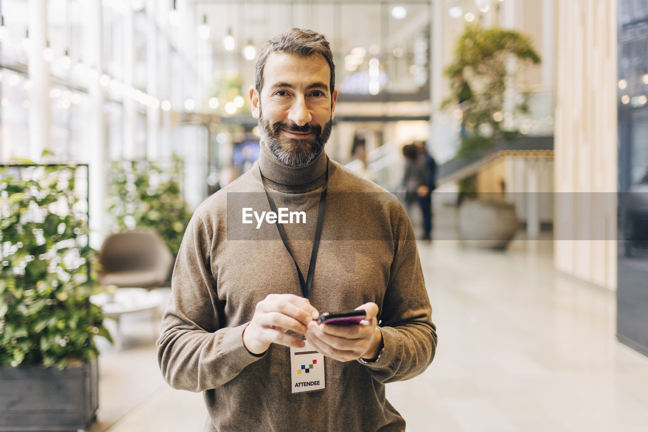 Portrait of smiling mature businessman holding smart phone while standing at convention center
