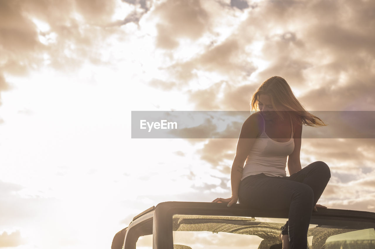 Low angle view of young woman sitting on car roof against sky during sunset