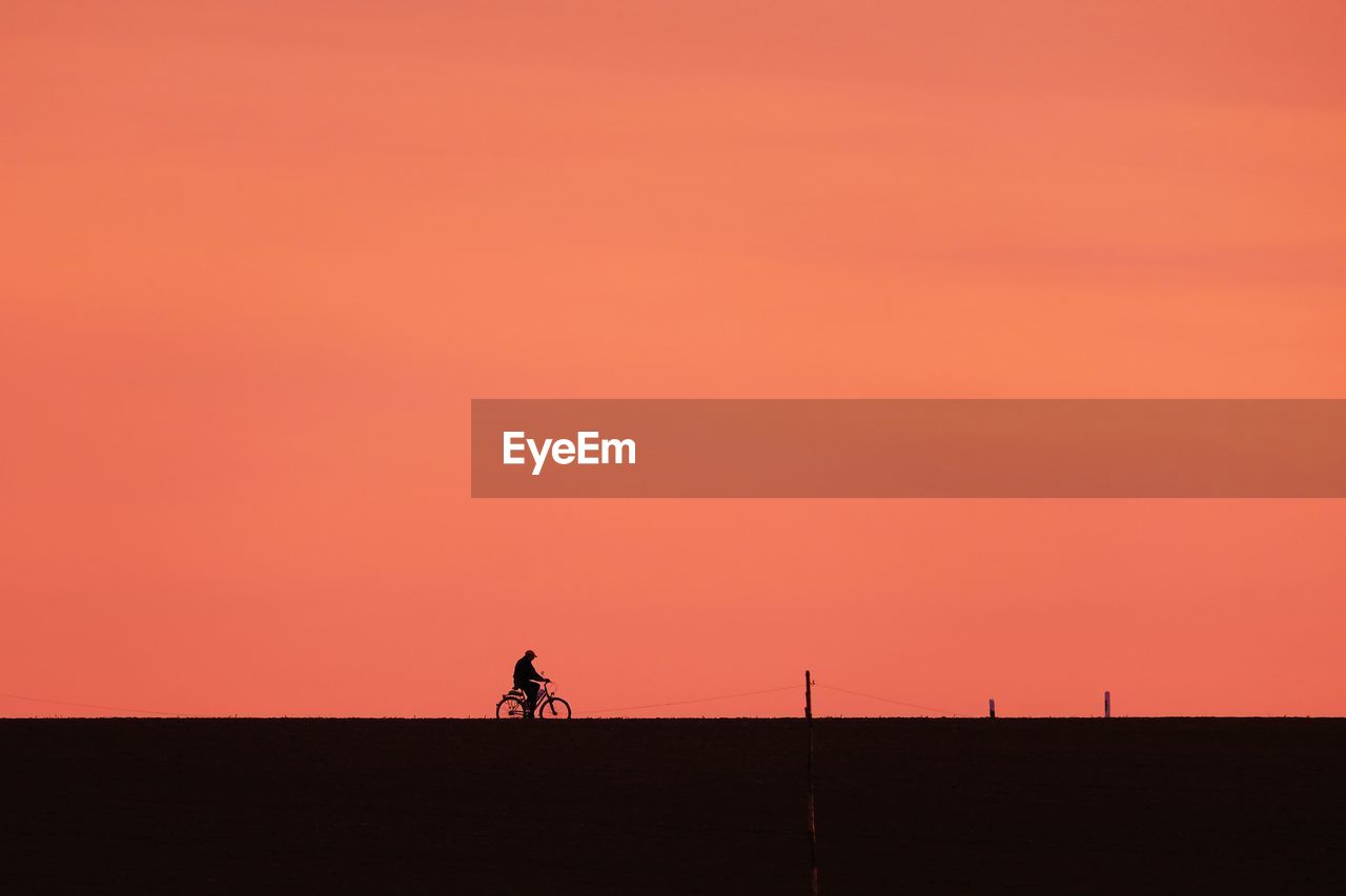 Silhouette man riding bicycle against orange sky