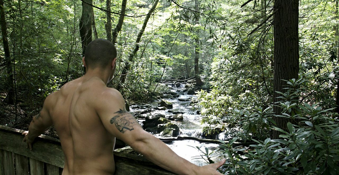 Rear view of shirtless muscular man looking at stream flowing in forest