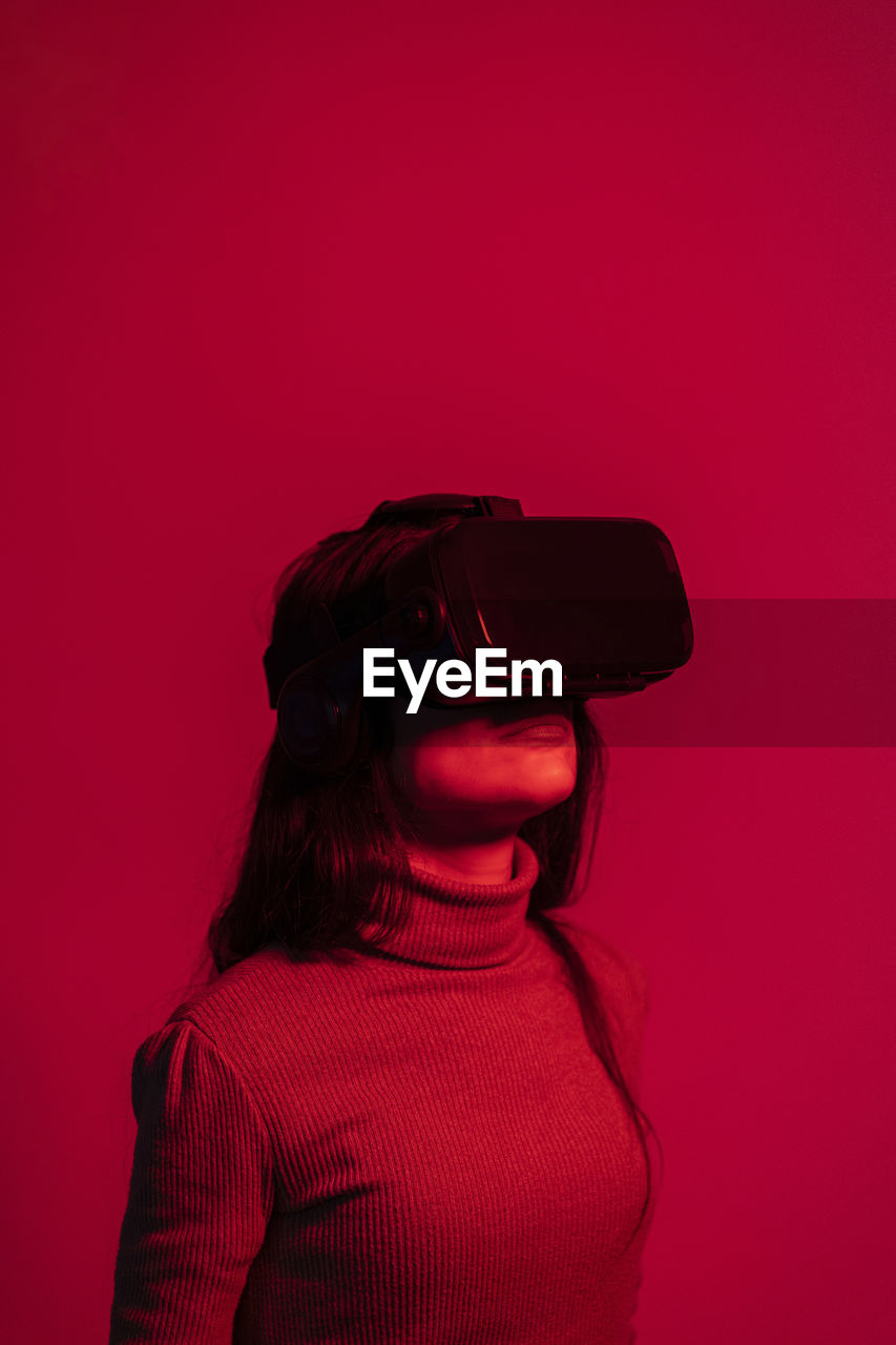 Mid adult woman using virtual reality headset while standing against red background