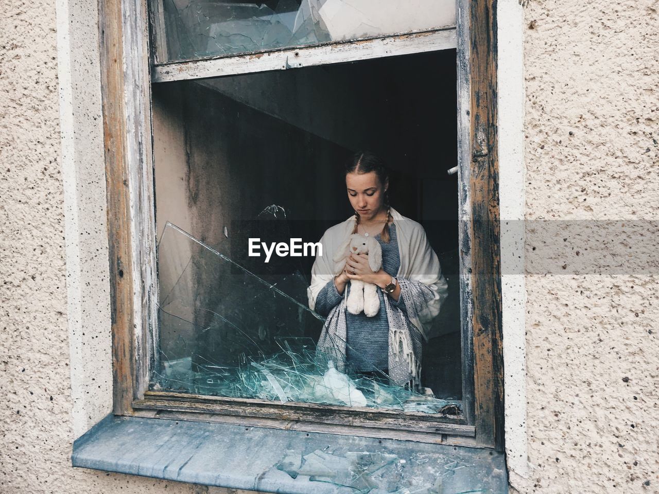 Woman with stuffed toy in building seen through broken window