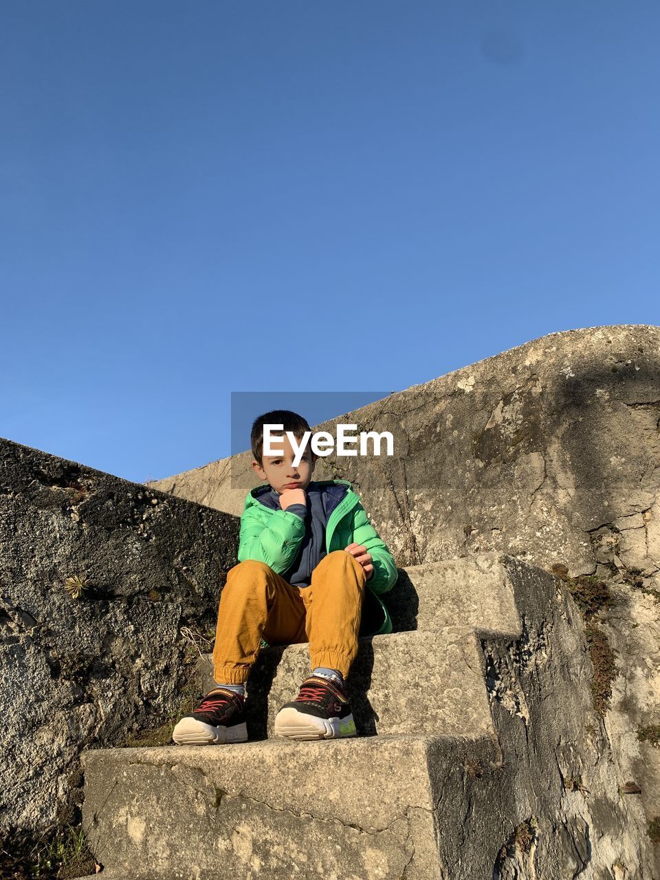 Full length of boy sitting on rock against clear blue sky