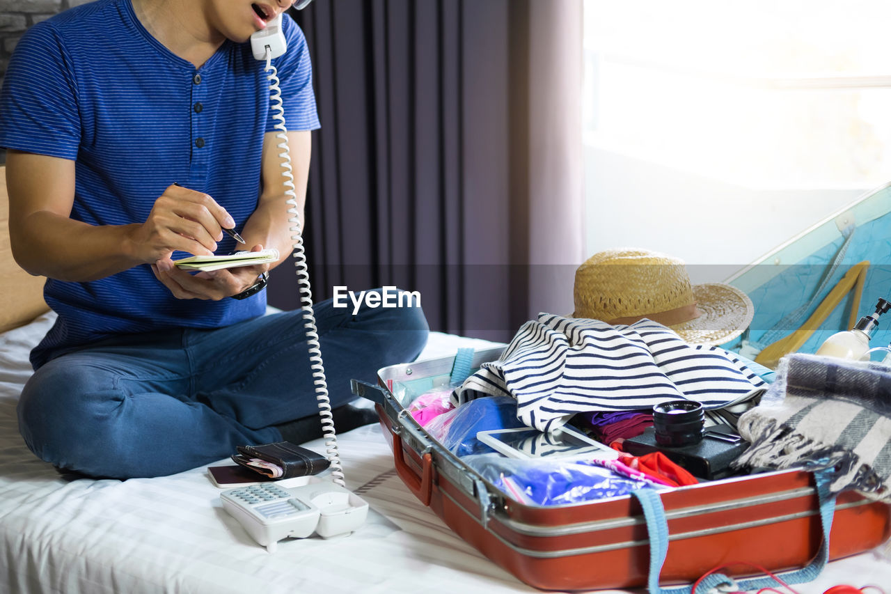 Low section of man using telephone while packing luggage on bed at home