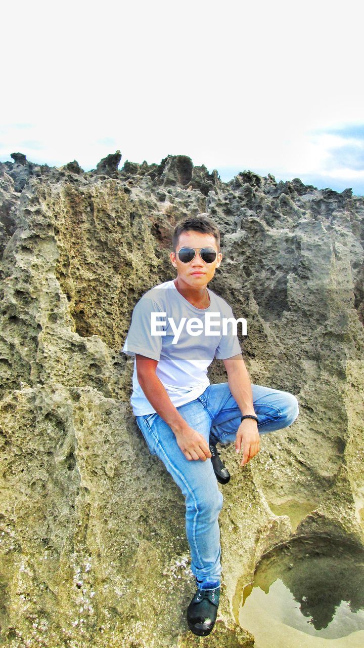 Portrait of man wearing sunglasses standing on rock against sky