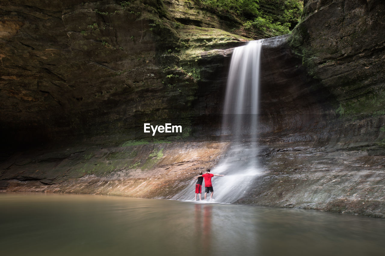 Rear view of boys standing on rock formation by waterfall in forest