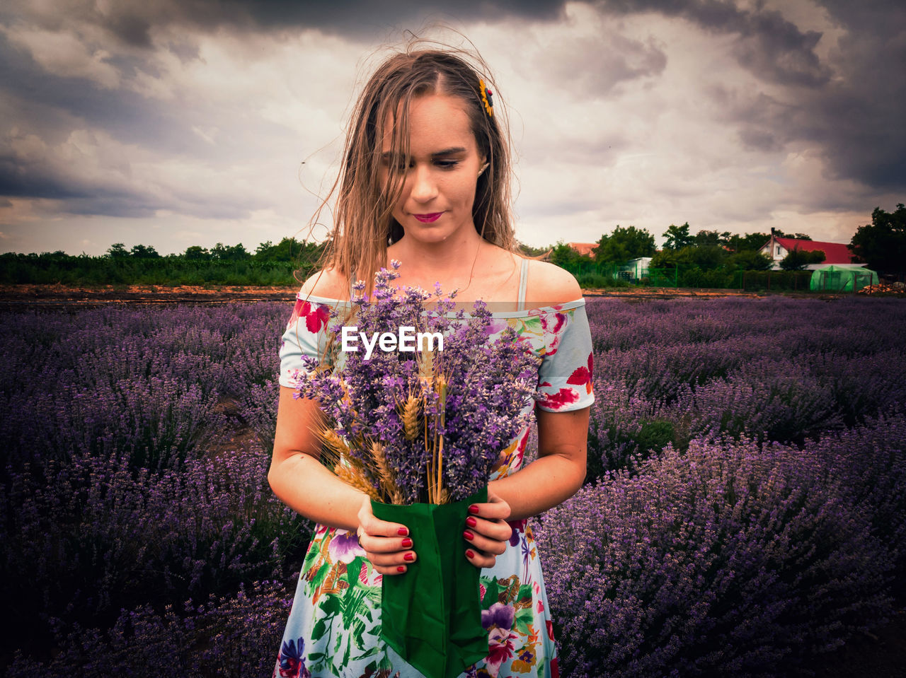 Woman holding lavender flowers while standing on field against cloudy sky