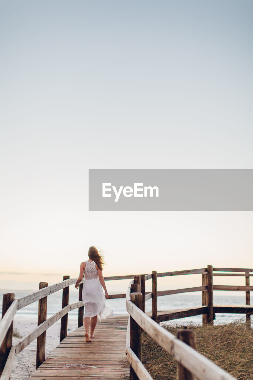 Rea view of young woman walking on pier at beach against clear sky during sunset