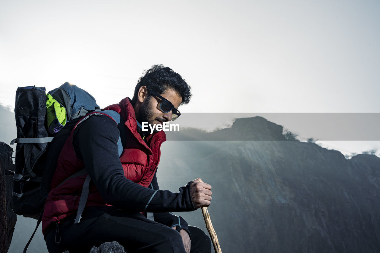 Young indian mountaineer sitting on the top of the mountain on a cliff with a backpack and a stick.