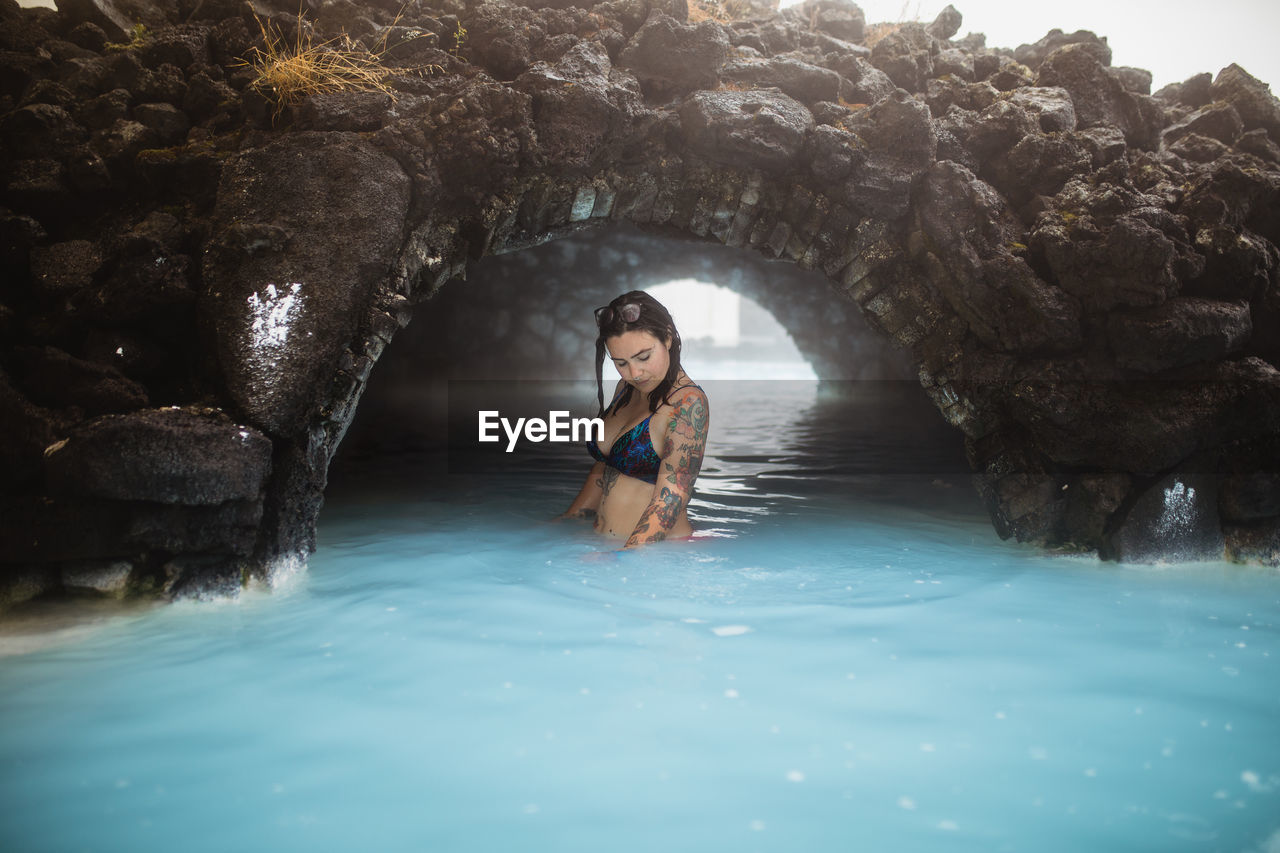 Side view of young tattooed hipster in swimsuit posing in blue water between rocks