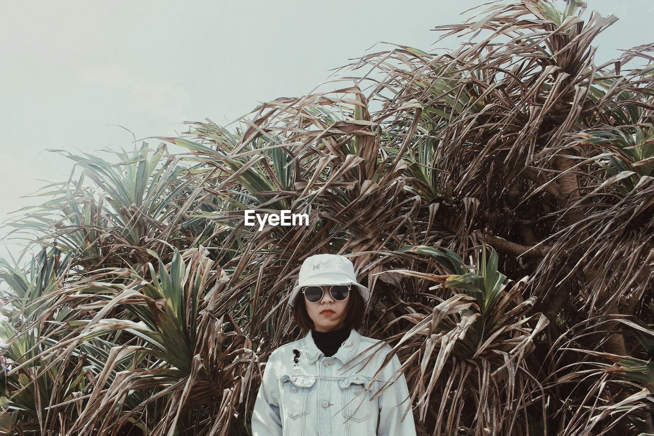 Portrait of young woman wearing hat and sunglasses standing against plants