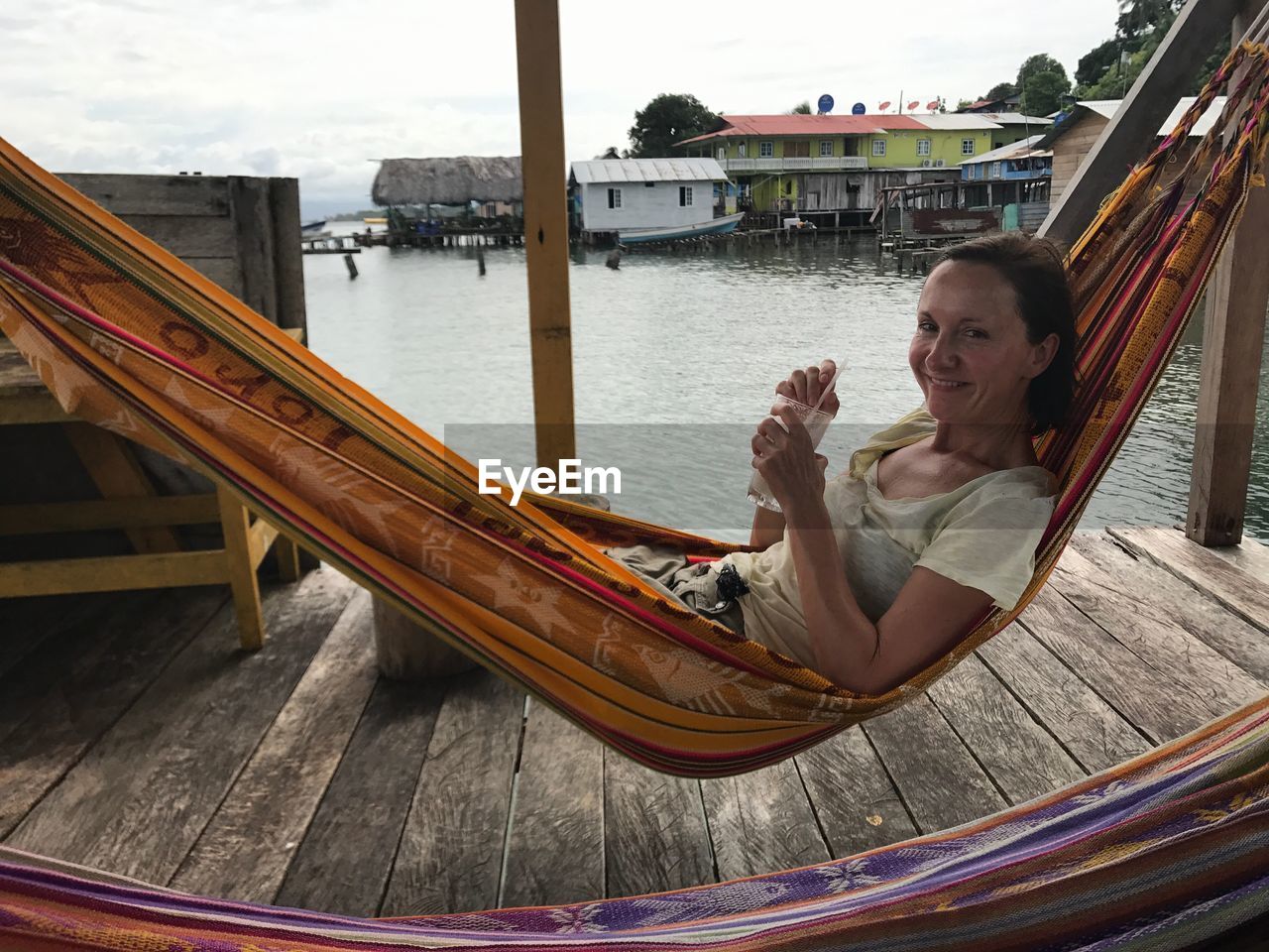 Portrait of smiling woman with drink resting in hammock by lake