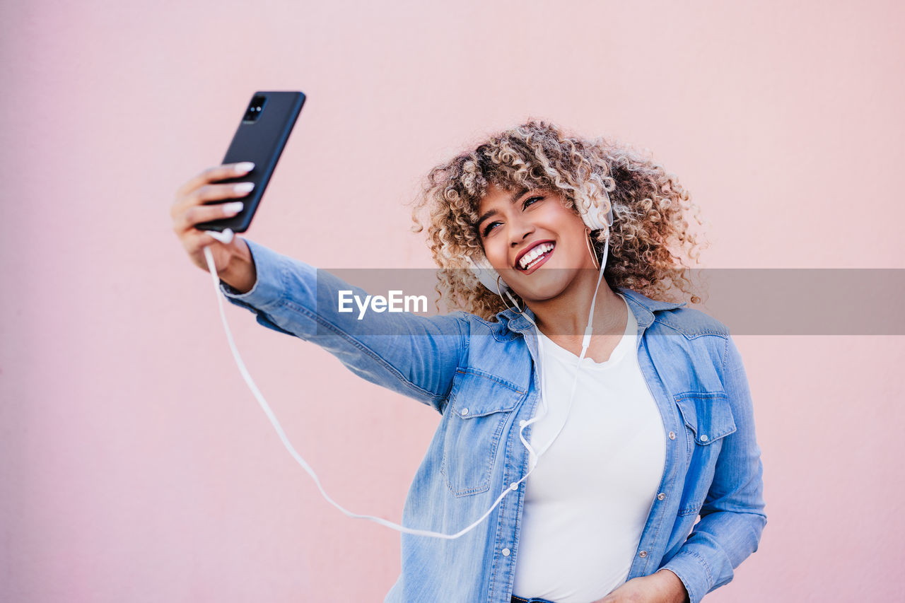 Portrait of smiling hispanic woman with afro hair in city using mobile phone and headset. lifestyle