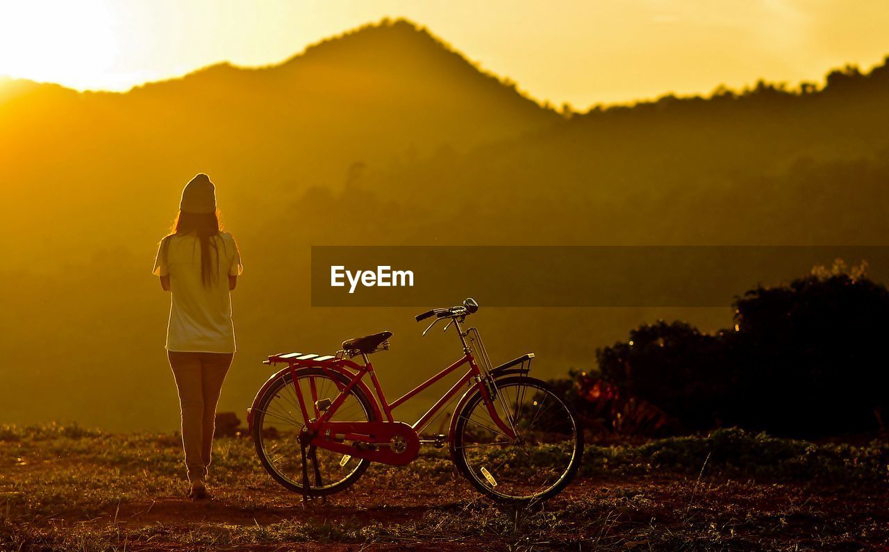 Woman walking by bicycle against mountains during sunset