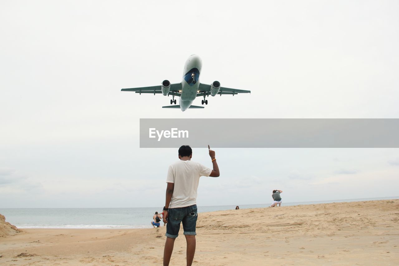 Rear view of man standing while pointing towards airplane at beach 