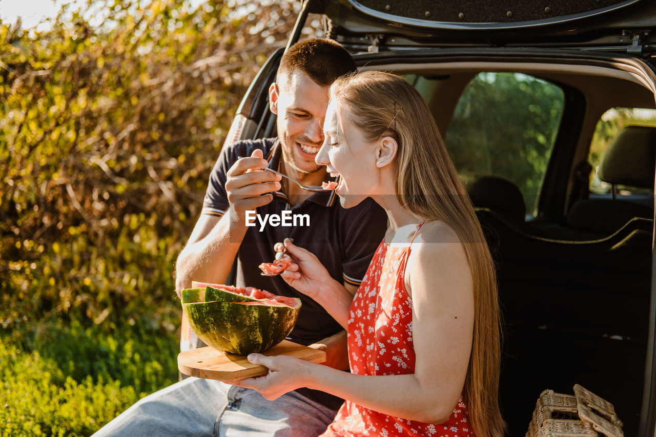 Millennial couple sitting on open trunk and eating watermelon. happy young couple having break at