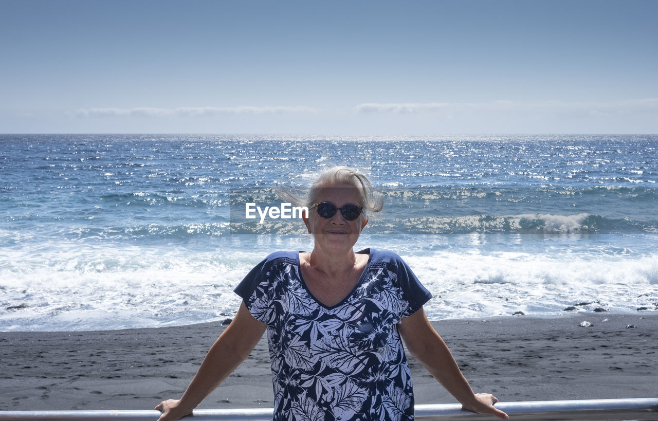 Portrait of senior woman wearing sunglasses on beach against sky