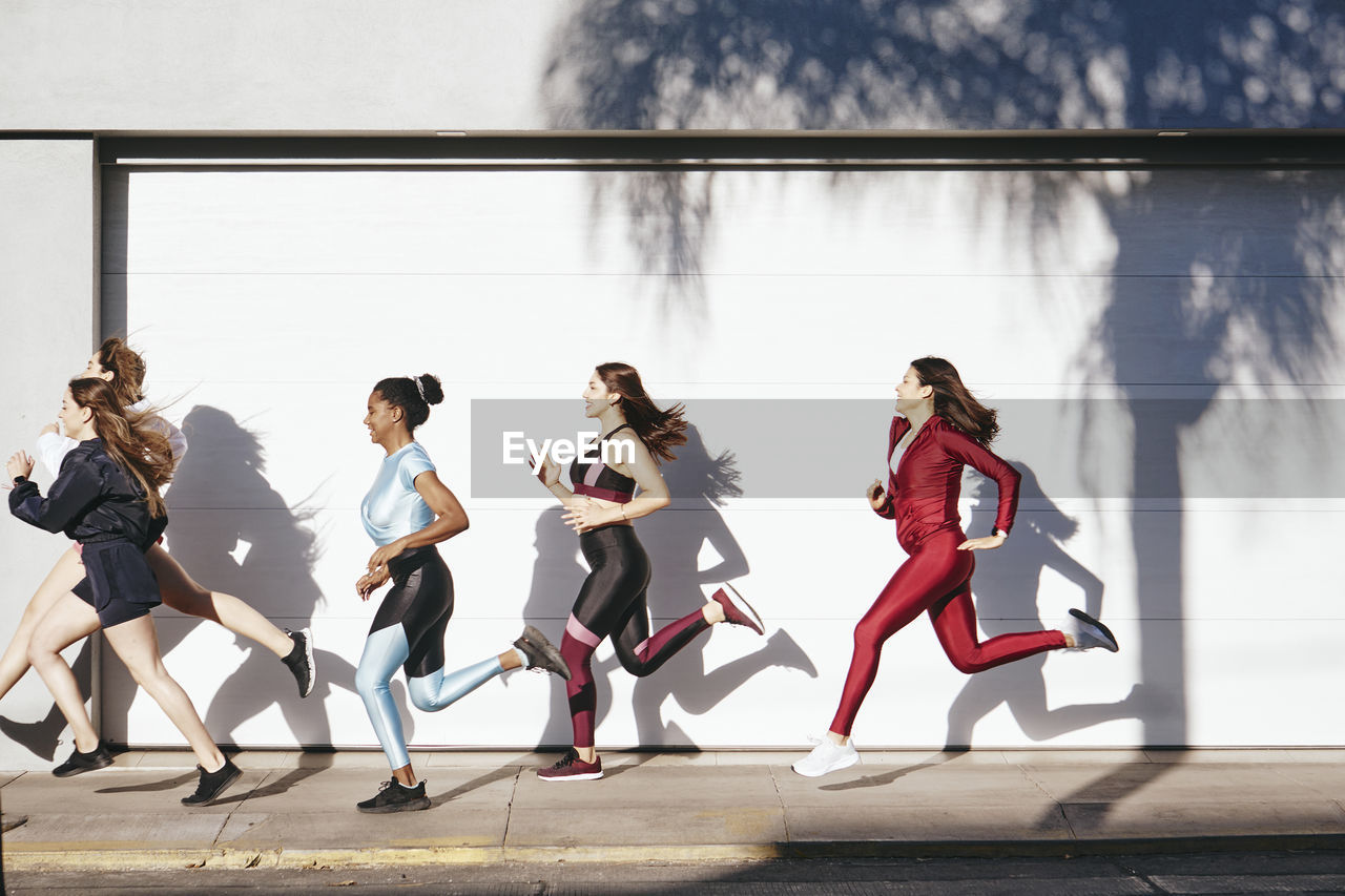 Side view full body of young fit multiracial female athletes in stylish sportswear running on pavement near white wall during outdoor fitness training on sunny day