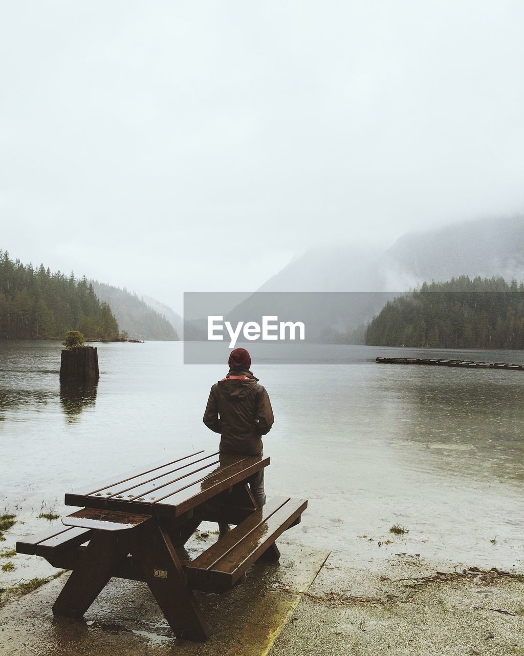 Rear view of man standing on lakeshore by bench against sky during foggy weather