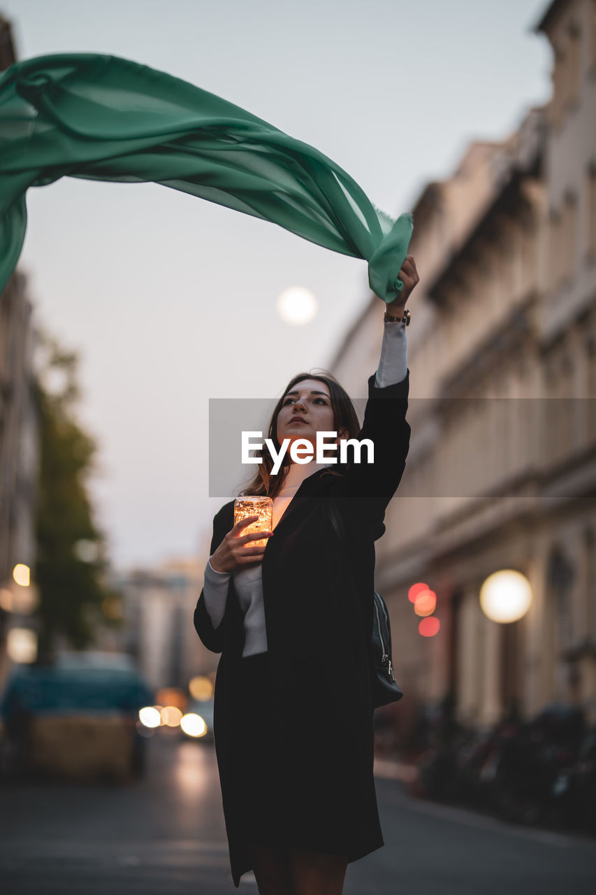 Woman moving green scarf while holding illuminated jar in city at dusk