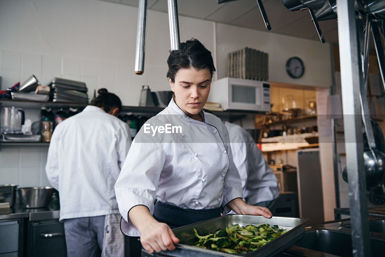 Mid adult female chef holding baking sheet with broccoli at commercial kitchen