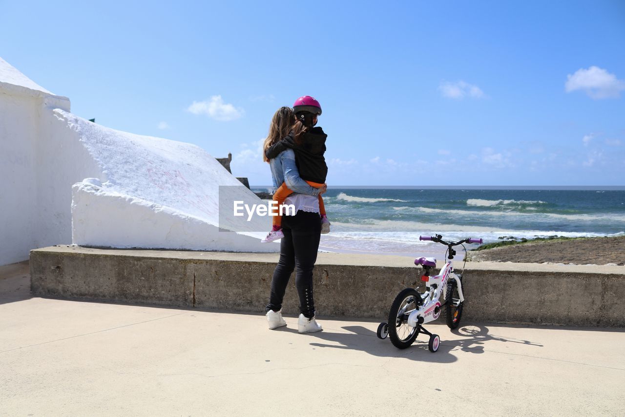 Rear view of mother carrying daughter while standing by bicycle against sea