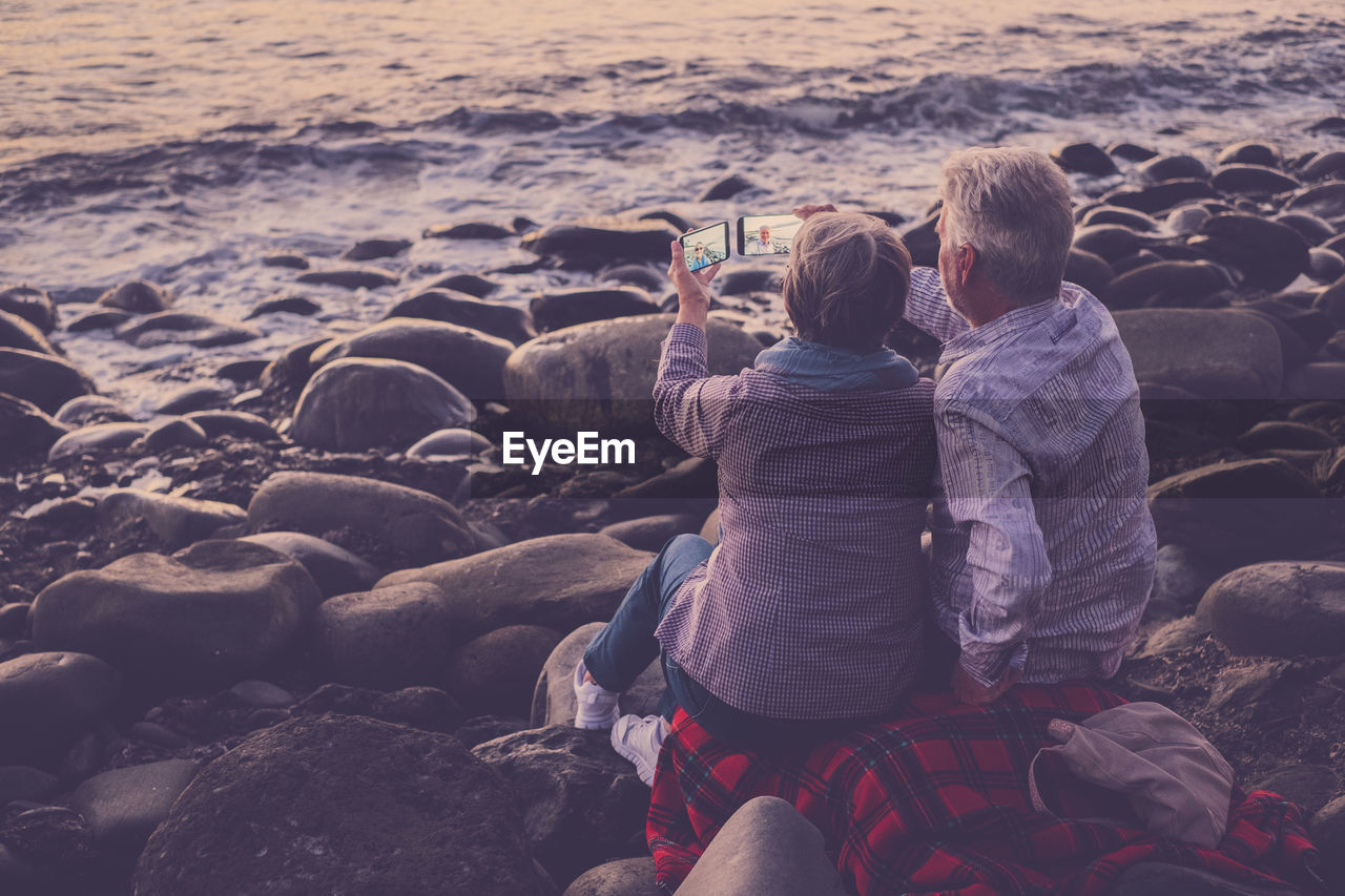 Rear view of couple taking selfie from mobile phones at beach