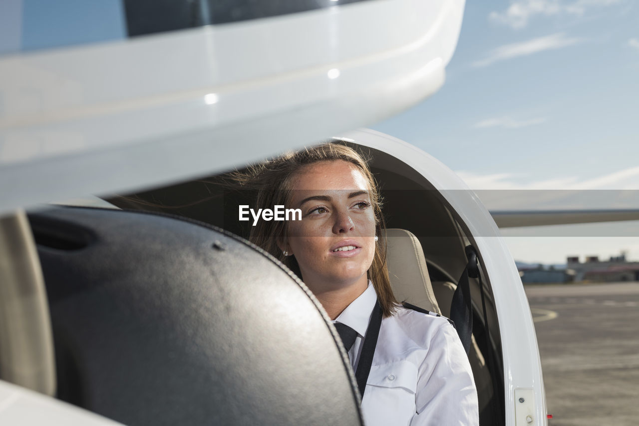 Thoughtful female pilot sitting in airplane against sky at airport