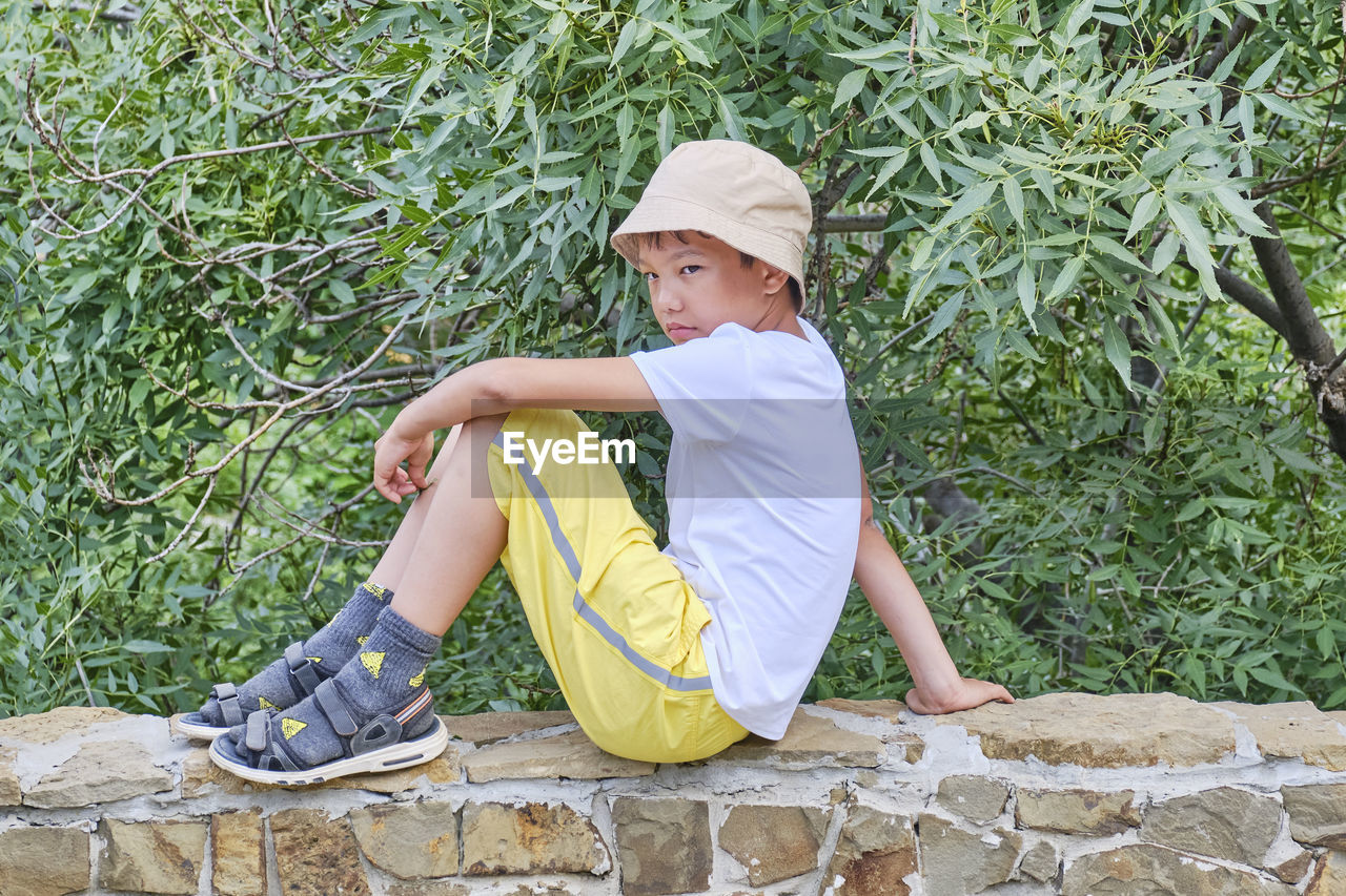 SIDE VIEW OF BOY SITTING ON STONE WALL