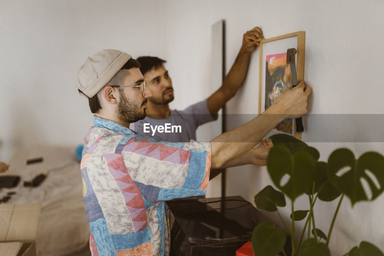 Side view of man hanging frame on wall with boyfriend at home