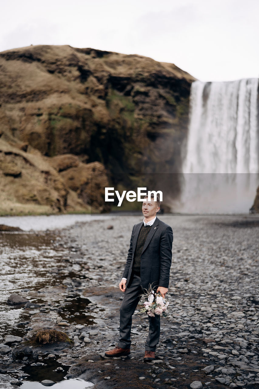 PORTRAIT OF MAN STANDING ON ROCK AGAINST WATERFALL