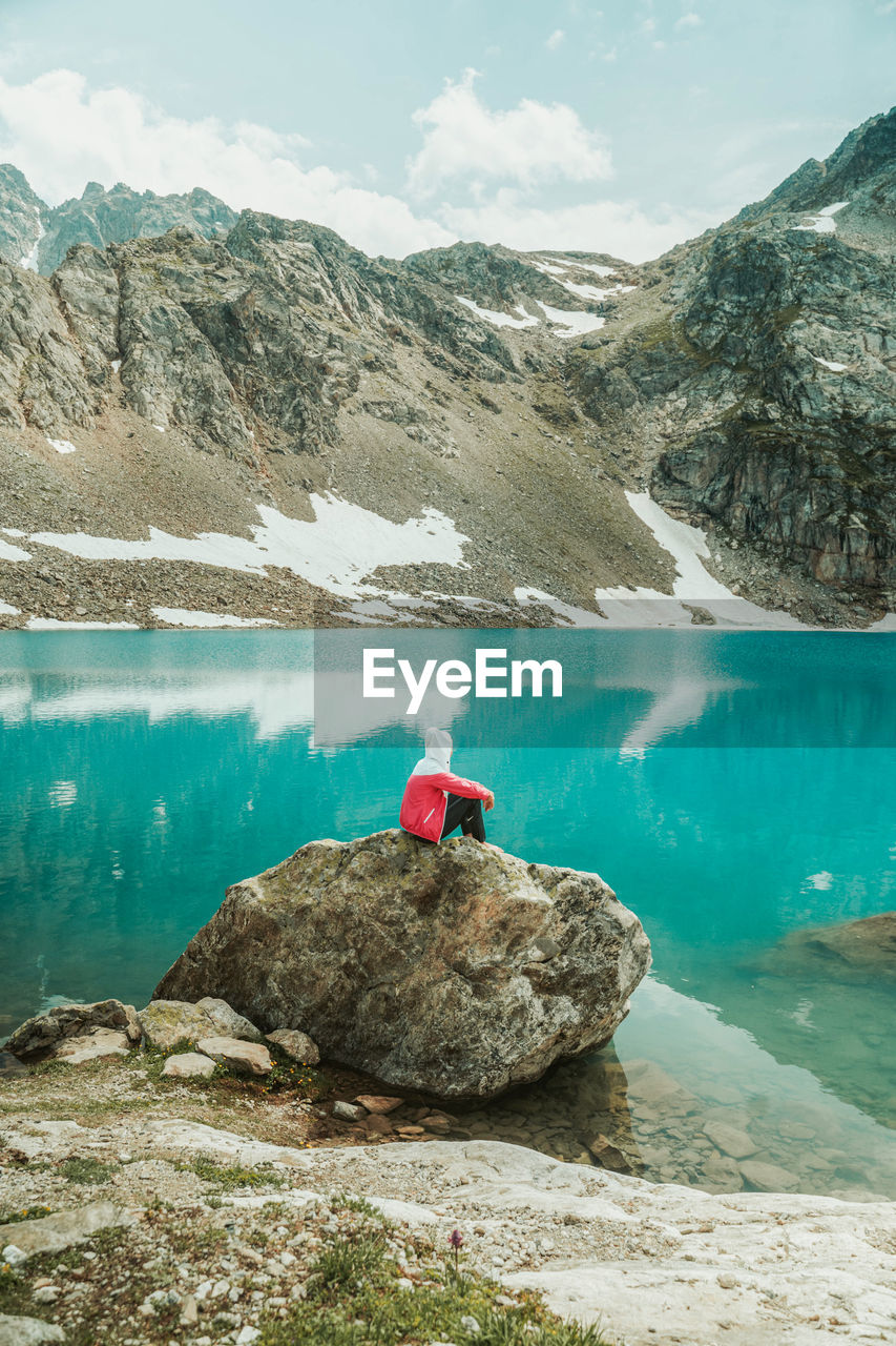 MAN ON ROCKS BY LAKE AGAINST MOUNTAIN