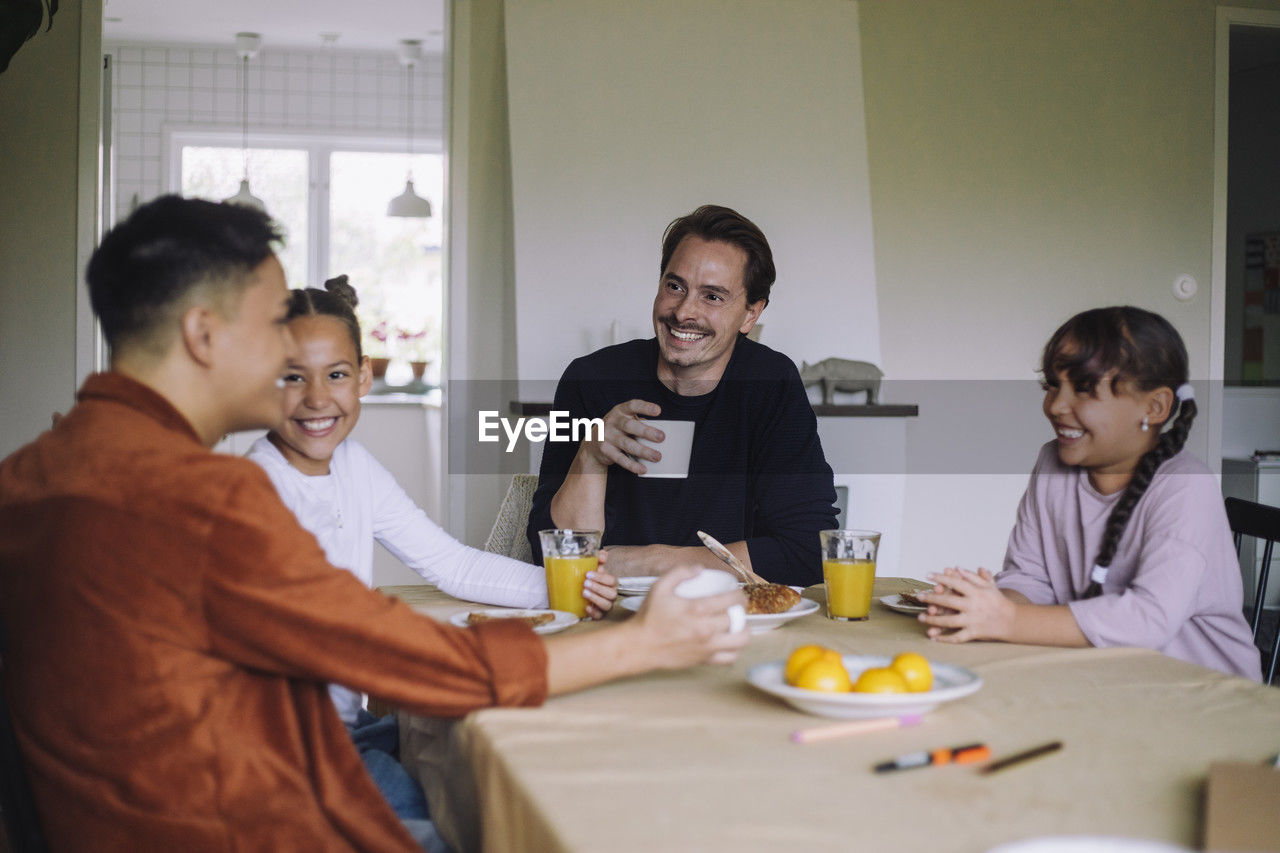 Happy gay couple spending leisure time with daughters while having breakfast at dining table in home