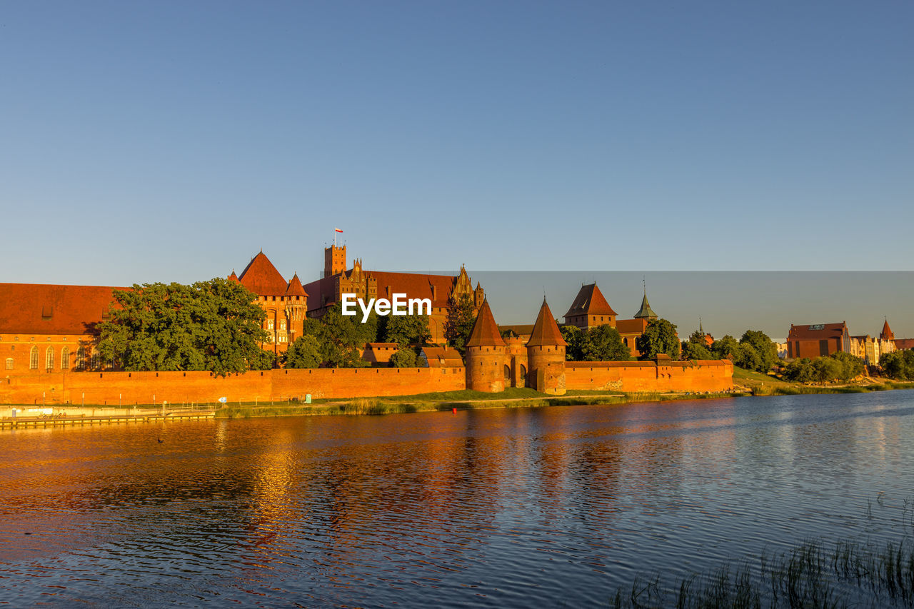 Buildings by lake against a clear blue sky, malbork castle. 
