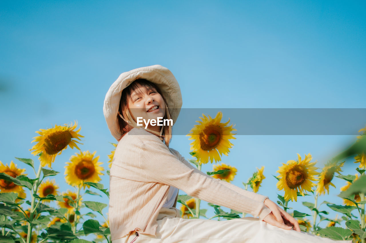 PORTRAIT OF SMILING YOUNG WOMAN WITH SUNFLOWER AGAINST PLANTS