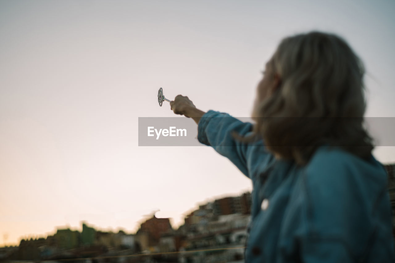 Side view of unrecognizable dreamy female sitting on roof of building and casting spell with magic wand in outstretched hand while looking away