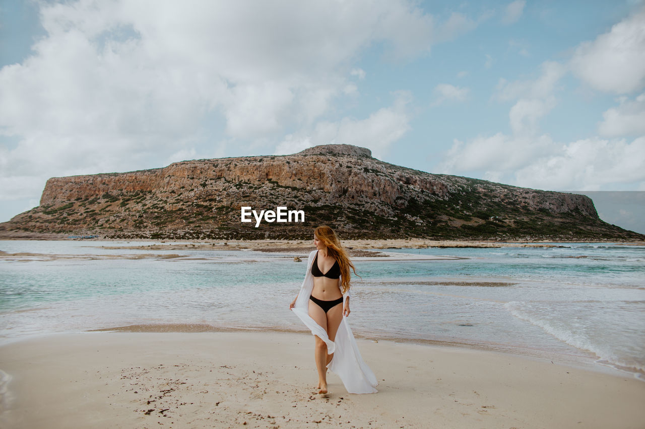 Full length slim female in black swimsuit standing on sandy balos beach against rocky cliff and looking away on clear sunny weather