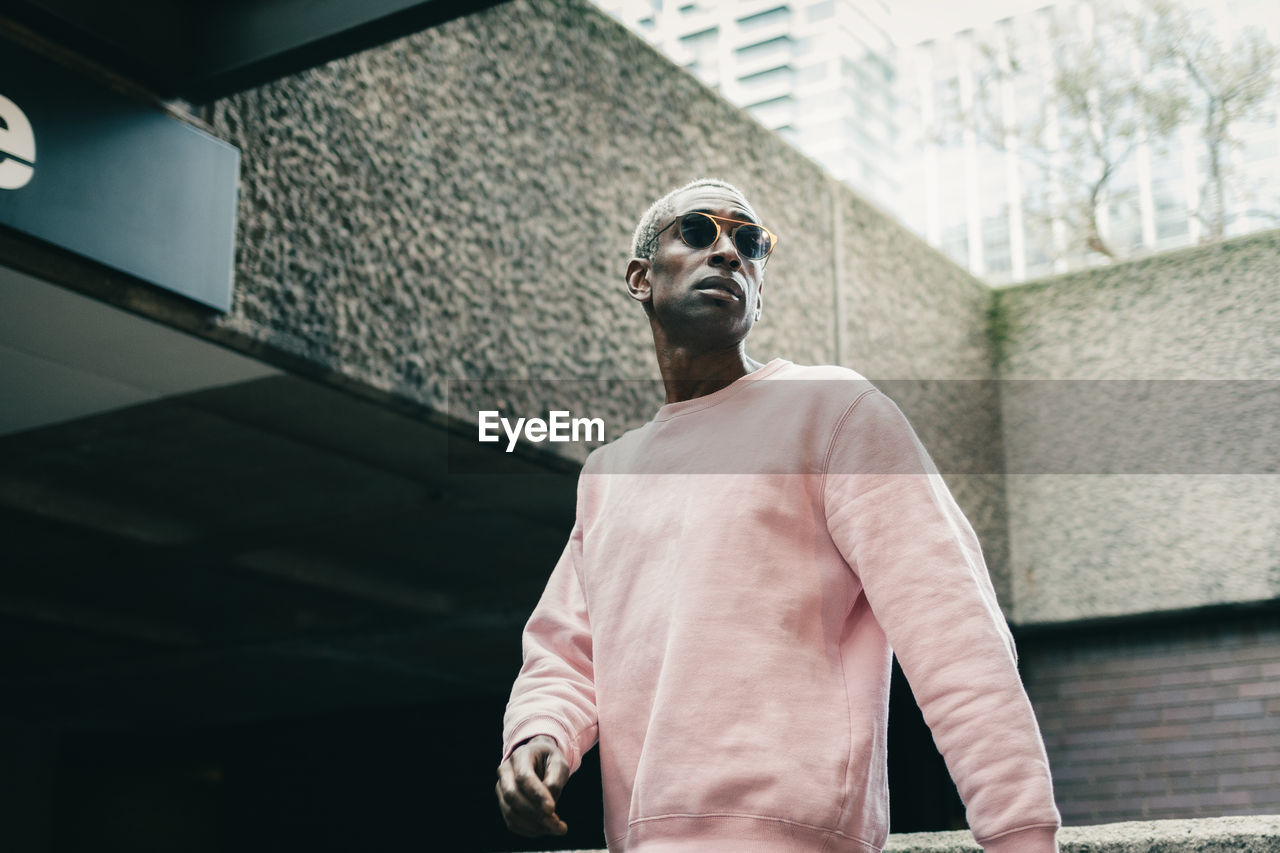 Low angle of confident black guy in stylish pink sweatshirt and sunglasses looking away and walking near underground entrance on city street