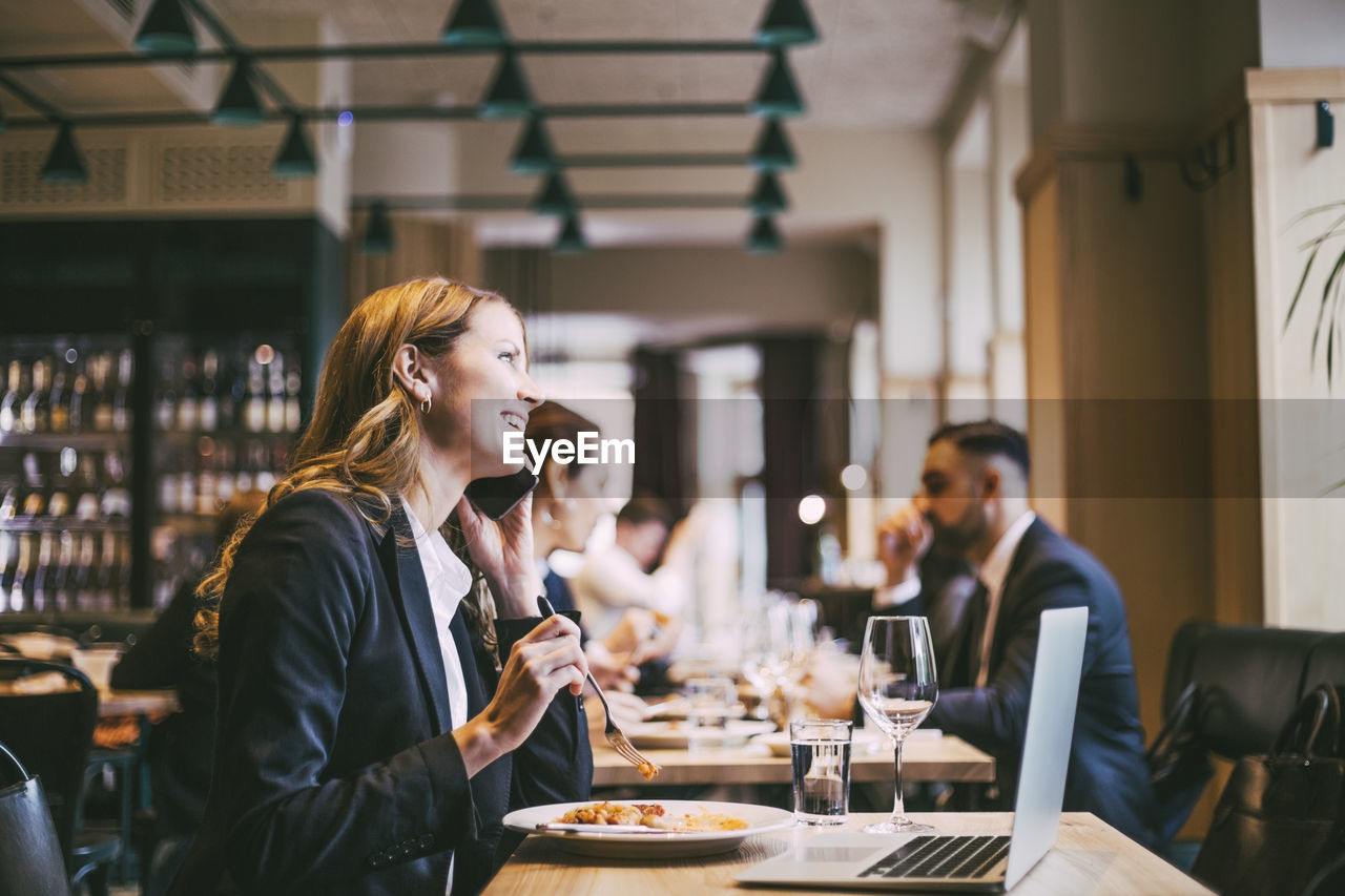 Smiling businesswoman talking on smart phone while eating in restaurant