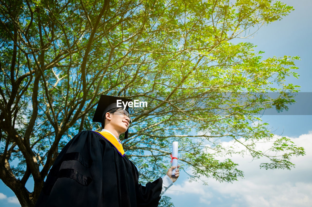 Young man in graduation gown standing against sky