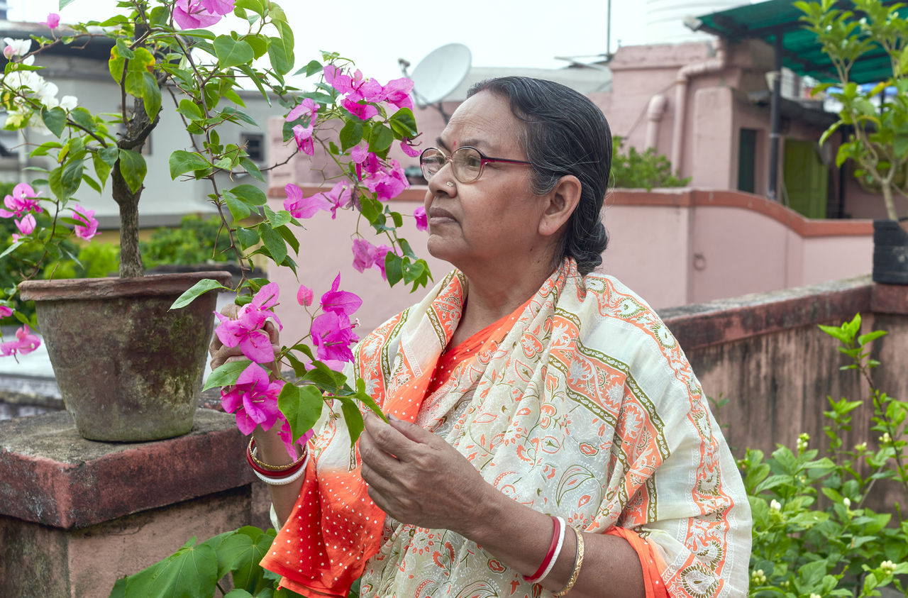 adult, women, one person, plant, nature, glasses, senior adult, lifestyles, flowering plant, flower, eyeglasses, female, mature adult, gardening, floristry, growth, seniors, holding, smiling, waist up, outdoors, looking, day, happiness, leisure activity, portrait, clothing, potted plant, architecture, standing, beauty in nature, emotion, front or back yard, garden, hairstyle, short hair, domestic life