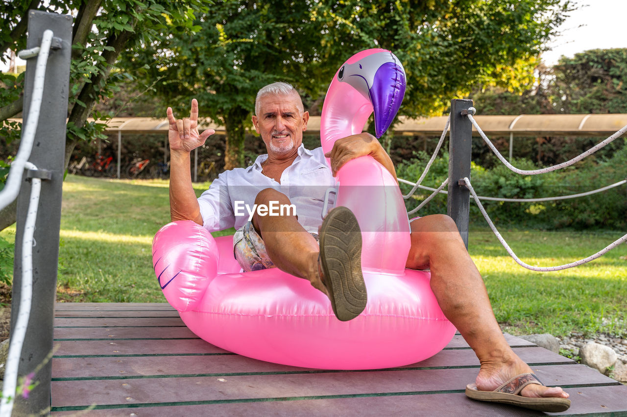 Smiling handsome middle aged man making sign of the horns sitting on an inflatable pink flamingo toy