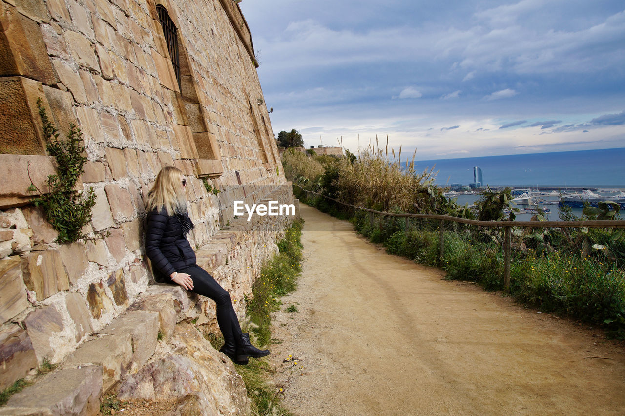 Side view of woman looking at sea against sky while sitting by wall