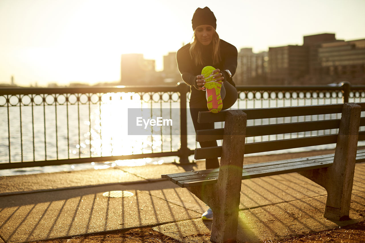 A backlit woman stretches pre-run on a park bench.