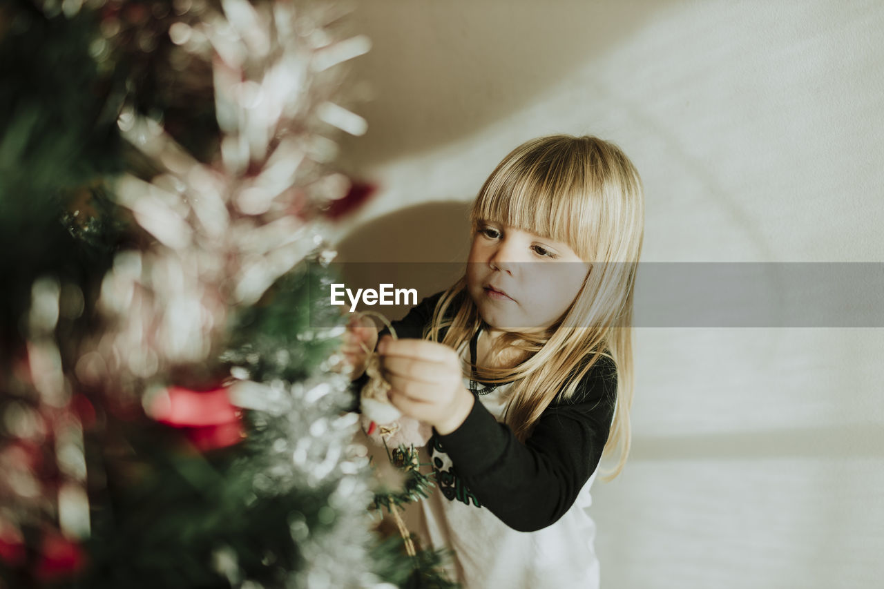 Blond girl decorating christmas tree against wall at home